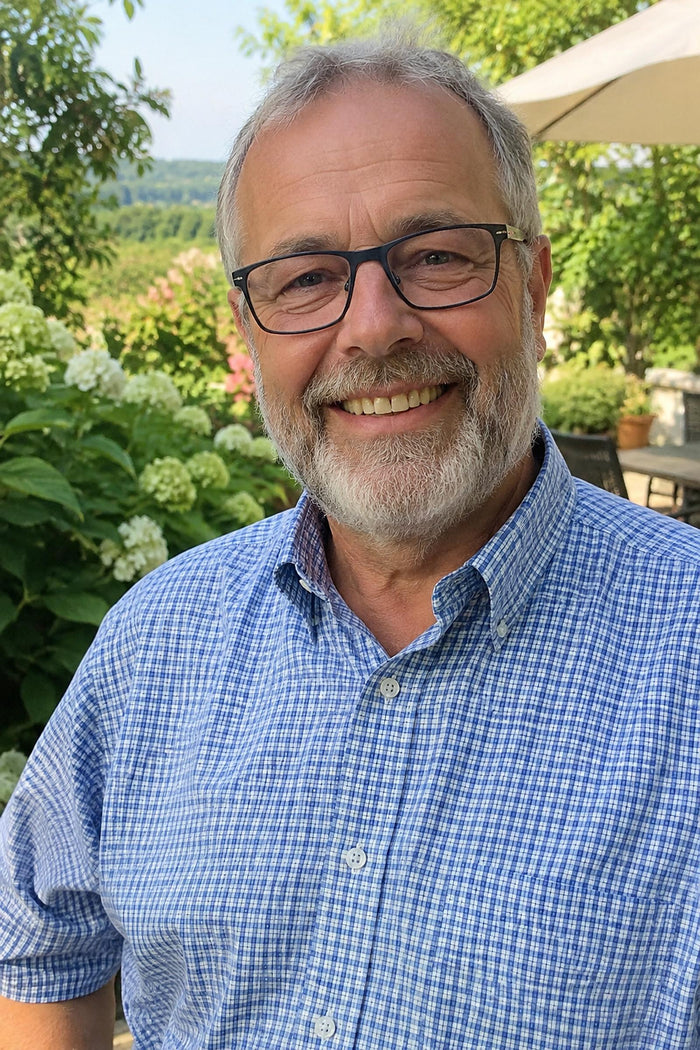 Ausgangsfoto: lächelnder Mann um die 65 mit grauem Vollbart, eckiger Brille und hellblau kariertem Hemd im Garten